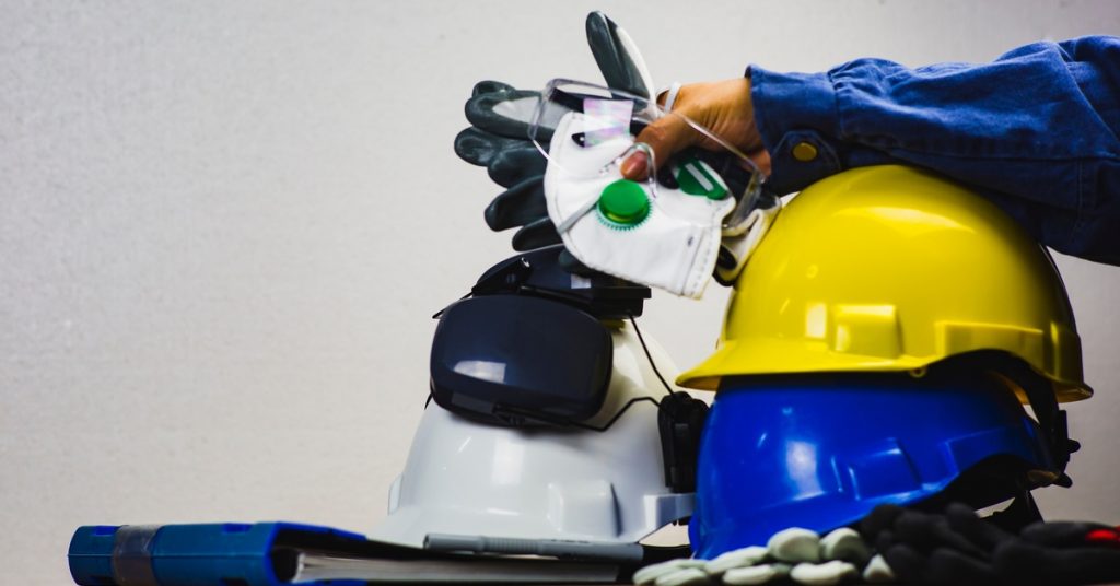 A person is holding some PPE, including goggles and gloves, while their arm rests on a stack of helmets.