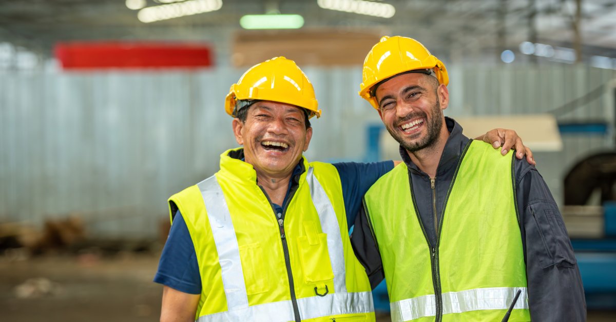 Two workers wearing high-visibility vests and yellow hard hats stand in a factory and smile at the camera.