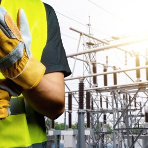 A close-up of someone securing protective gray-and-yellow gloves on their hands. The person stands near a power station.