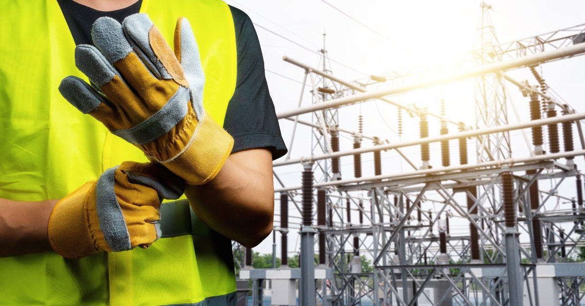 A close-up of someone securing protective gray-and-yellow gloves on their hands. The person stands near a power station.