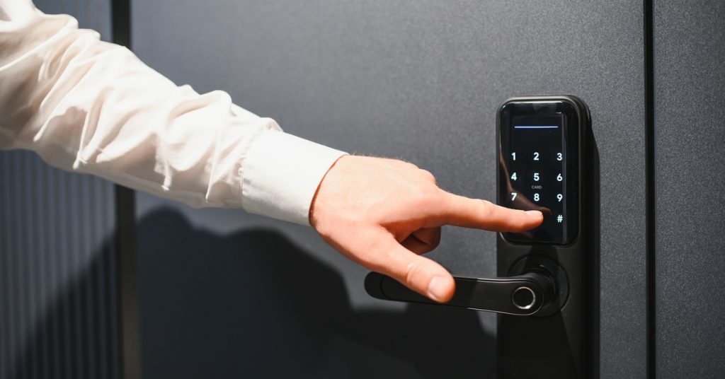 A close-up of a man's hand touching a digital panel on a door security system. He wears a white shirt.