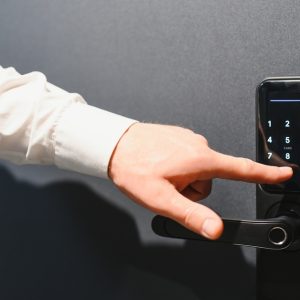 A close-up of a man's hand touching a digital panel on a door security system. He wears a white shirt.