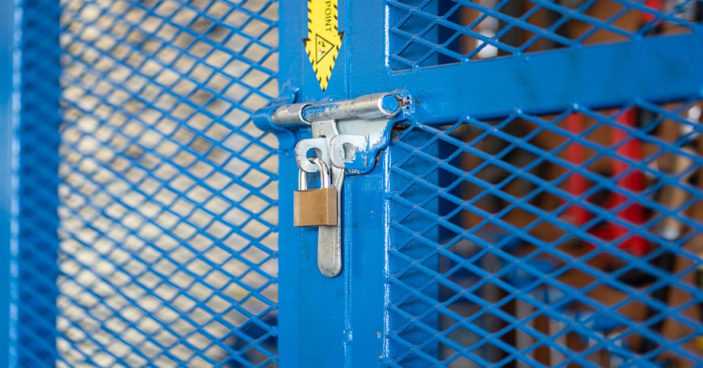 A close-up of a keyed padlock on a blue fenced gate. There is a yellow and black arrow sticker pointing toward the lock.