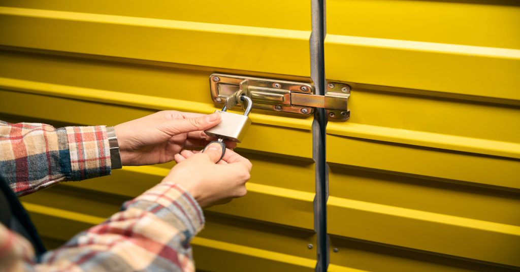 A close-up of a person in a plaid shirt unlocking a padlock on a yellow metal storage unit door with a key.