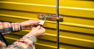 A close-up of a person in a plaid shirt unlocking a padlock on a yellow metal storage unit door with a key.