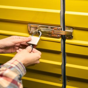 A close-up of a person in a plaid shirt unlocking a padlock on a yellow metal storage unit door with a key.