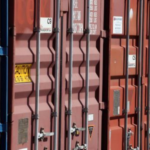 A row of red and blue shipping containers with vertical steel locks. Sunlight shines on the containers.