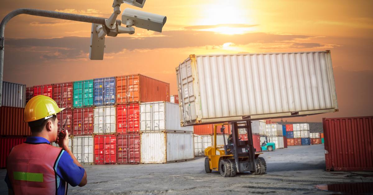 A shipping container yard with surveillance cameras. A worker has a walkie-talkie and looks at a machine lifting a container.