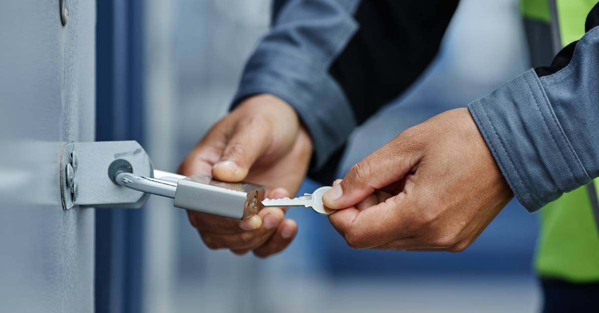 A close-up of a worker inserting a key into a padlock attached to a shipping container. The lock and container are gray.