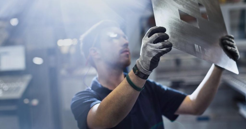 A man holds and examines a piece of steel while wearing gray and black safety gloves. The sun shines over his face.