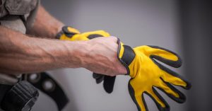 A close-up of a man with veiny arms and hands putting on yellow safety gloves. He's in front of a gray wall.