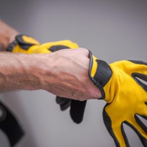 A close-up of a man with veiny arms and hands putting on yellow safety gloves. He's in front of a gray wall.