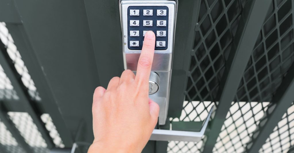 A close-up of a hand entering a code on a keypad lock mounted on a black iron gate. The lock also has a handle.
