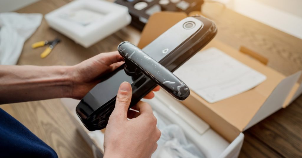 Someone holds a keyless door lock with the lock's packaging on the table below. Other packaging components lay on the side.