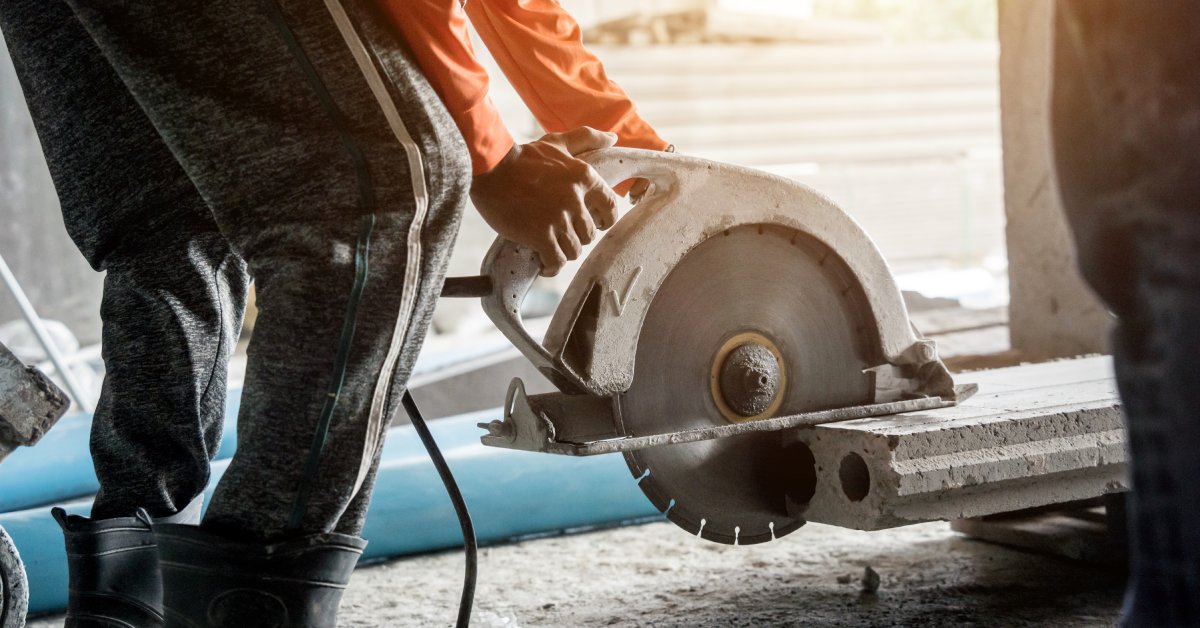 A worker wearing an orange shirt and long pants uses a large circular saw to cut a large piece of concrete.