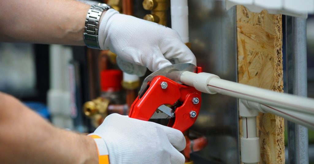 A worker wearing white gloves uses a red pipe cutter to cut a small plastic pipe. He has a watch on his left arm.