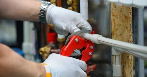 A worker wearing white gloves uses a red pipe cutter to cut a small plastic pipe. He has a watch on his left arm.