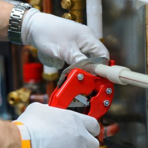 A worker wearing white gloves uses a red pipe cutter to cut a small plastic pipe. He has a watch on his left arm.