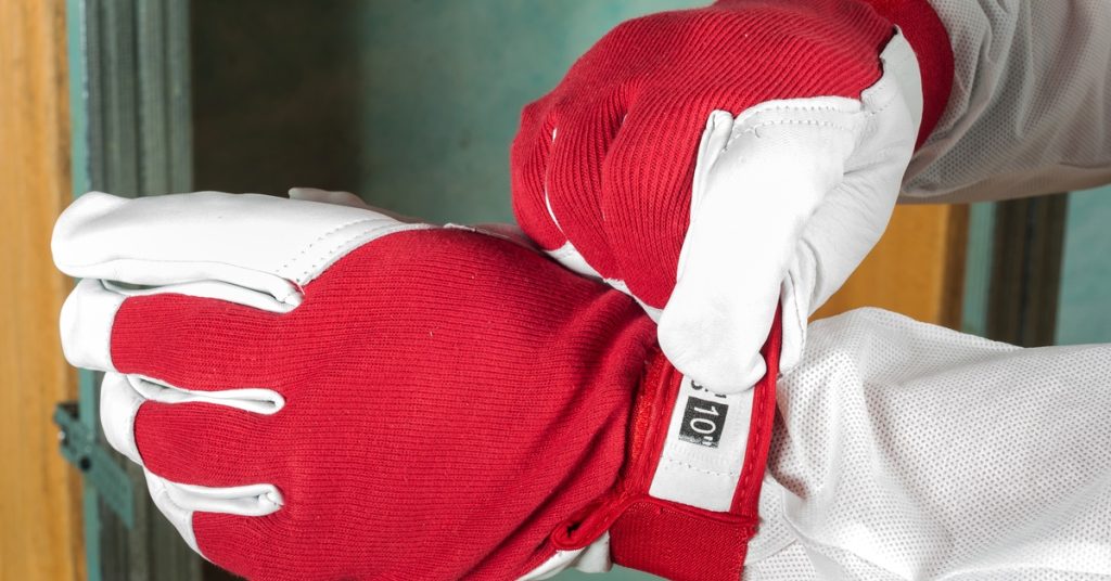 Red and white thermal gloves with ribbed cuffs and a size label worn by a worker installing insulation inside a home.