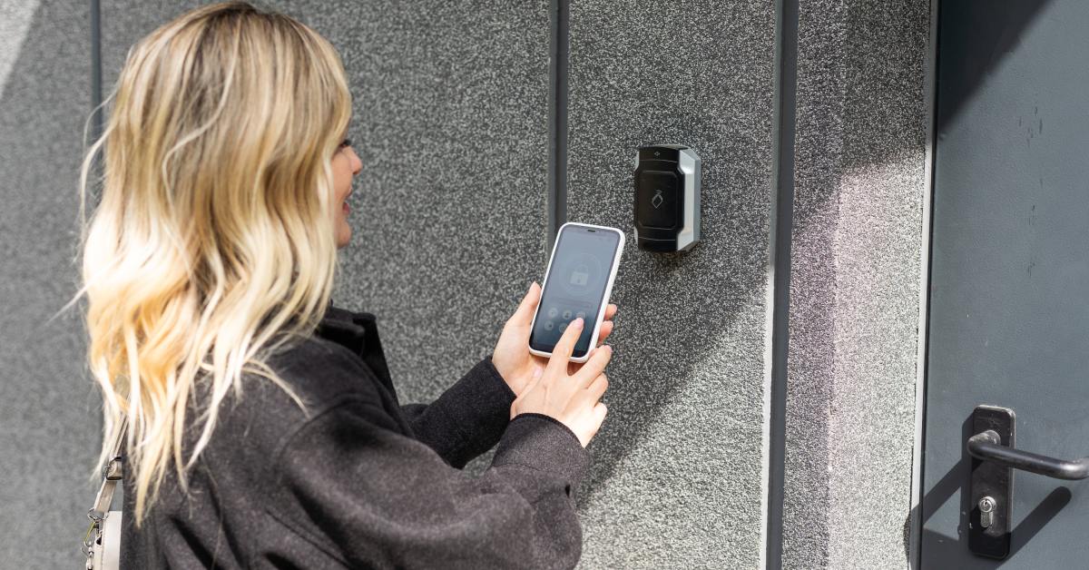 A young woman with blond hair uses her smartphone to unlock a controlled access panel outside of her apartment door.