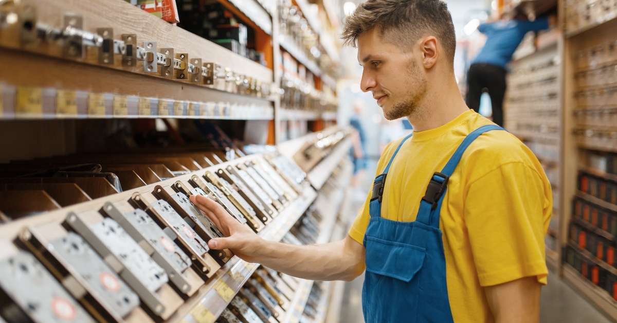 A man in a yellow shirt and blue overalls examines door locks and hardware on a display shelf in an industrial supply store.