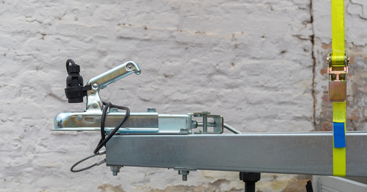 A trailer tow hitch coupler and wiring on a metal frame, with a yellow ratchet strap against a light brick wall.
