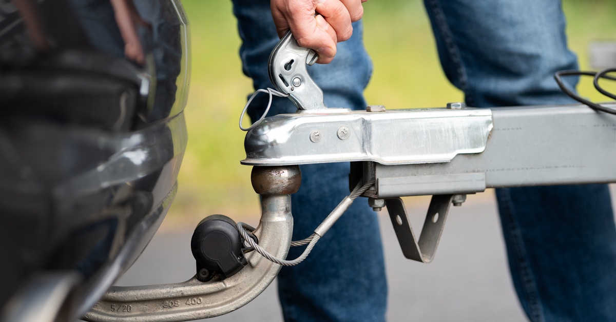 Close-up of a person attaching a trailer coupler to a car tow hitch, securing the latch and safety cable.