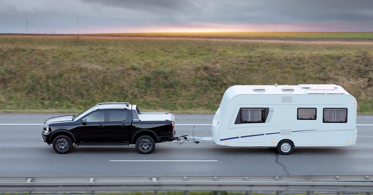 Black pickup truck towing a white travel trailer on a highway, passing open fields beneath a dark, cloudy sky.