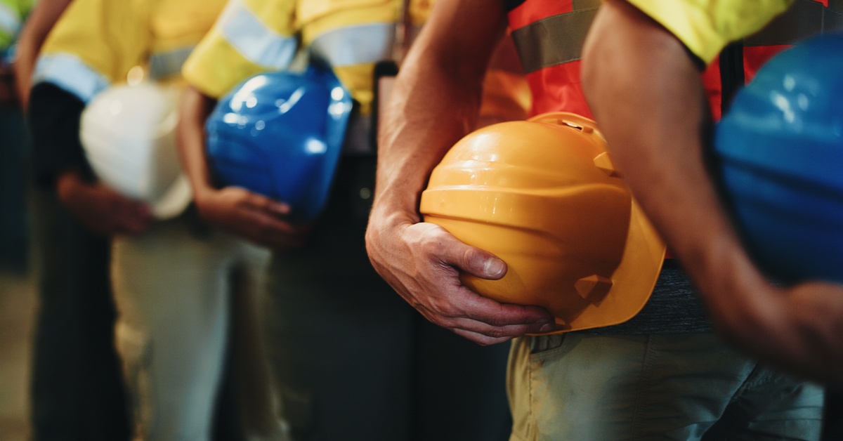 A group of construction workers wear safety vests and hold hard hats while standing together at a jobsite safety briefing.
