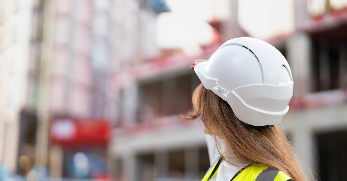 A construction worker wears a white hard hat and high-visibility safety vest while inspecting a building construction site.