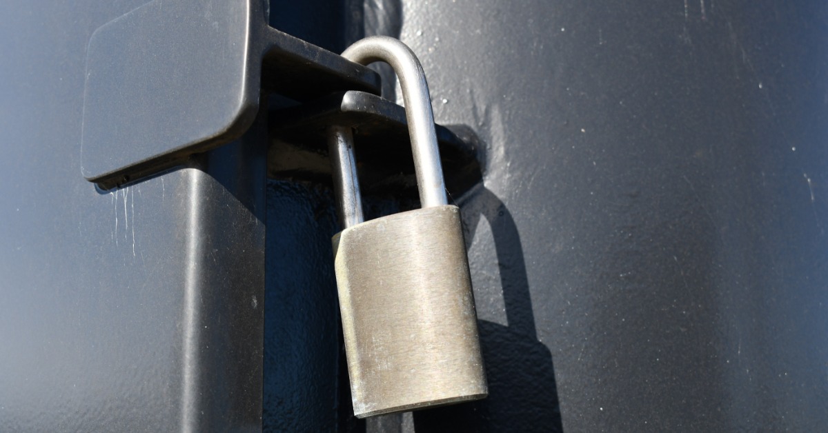 Close-up of a silver metal padlock securing a latch on a dark gray door or panel in bright sunlight.