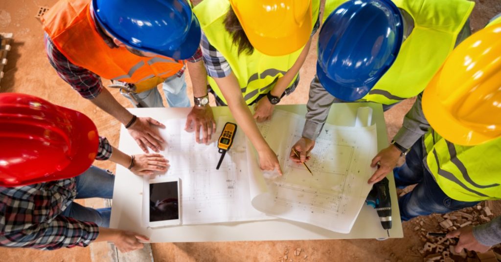 An overhead view of a construction team in colorful, non-vented hard hats, standing around a table to review blueprints.
