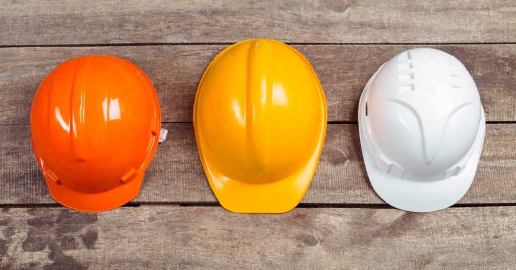 Three construction hard hats in orange, yellow, and white are arranged in a row on a wooden surface.