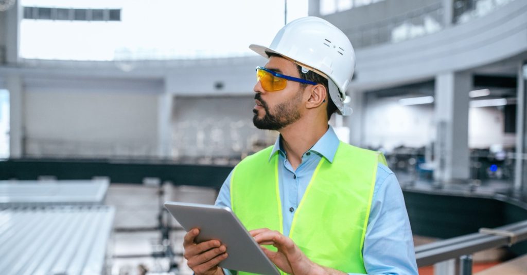 An industrial worker in a ventilated white hard hat and tinted safety glasses. He holds a tablet and looks off to the side.