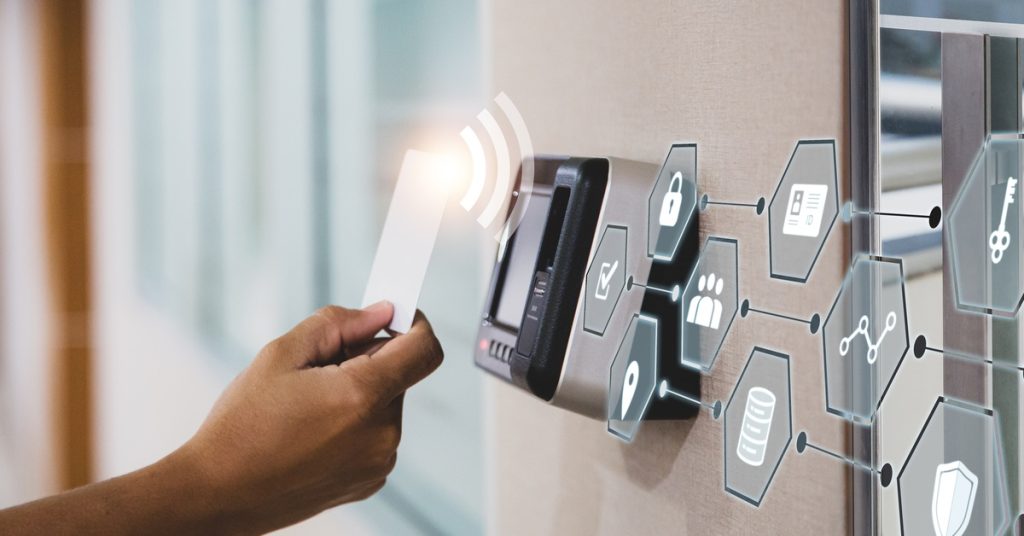 A hand taps a key card on a commercial access control system reader mounted on a wall next to a glass door.