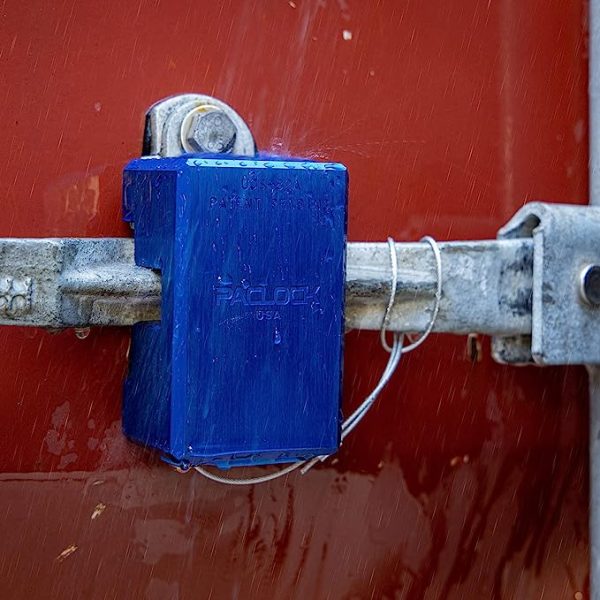 A large. blue, scuffed up locking mechanism keeping a metal latch locked and closed on the side of an orange container.