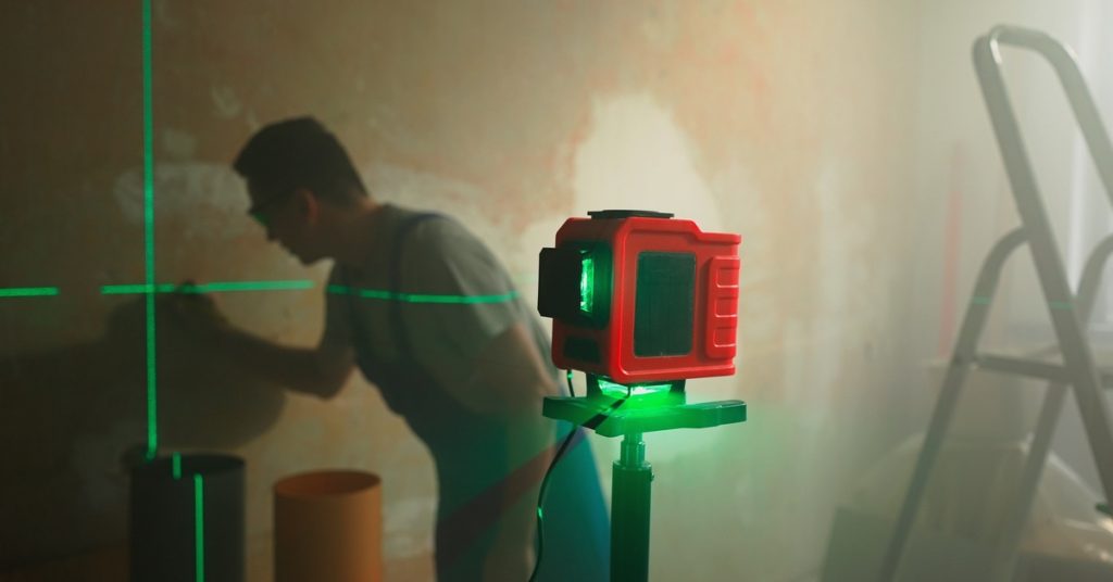 Green cross-line laser level on a tripod projecting beams onto a wall while a worker measures in the background.