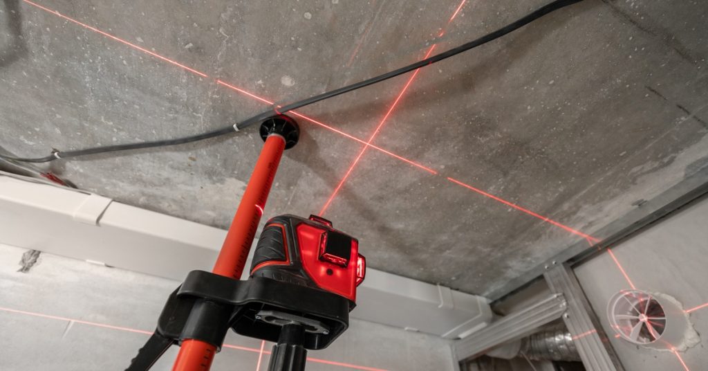 Red cross-line laser level mounted on a tripod projecting beams across a concrete ceiling at a construction site.