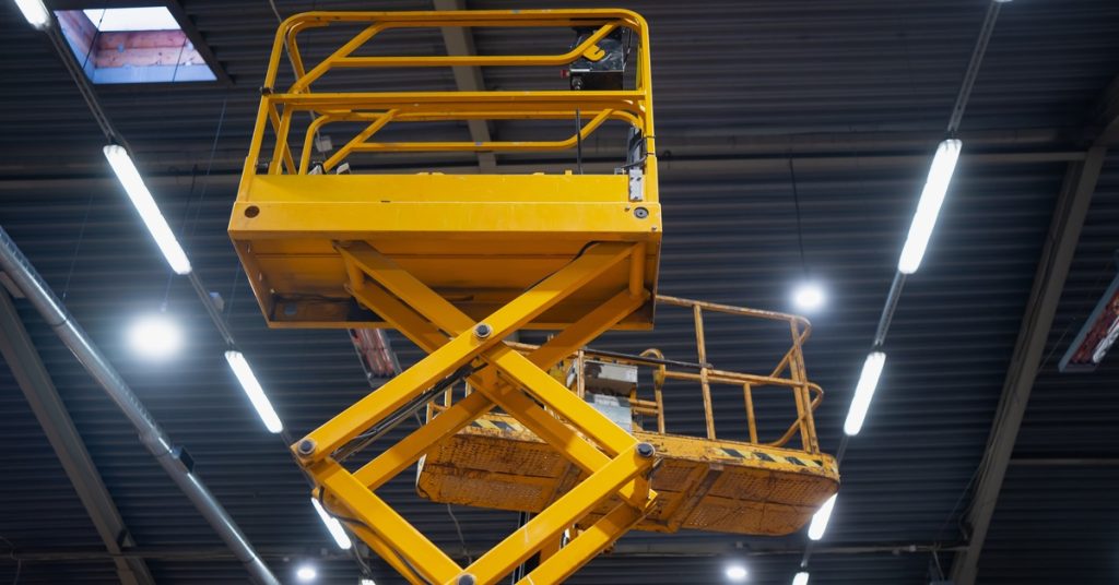 A yellow scissor lift raised inside a warehouse beneath bright overhead lights, exposed ceiling beams, and skylights.