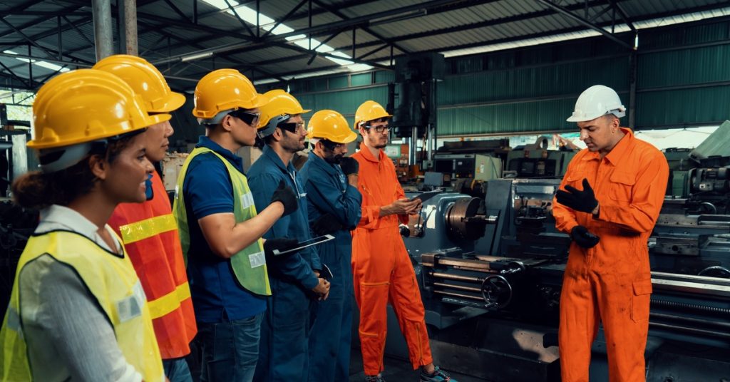 A group of workers in hard hats listen to a safety briefing beside industrial machinery inside a manufacturing facility.