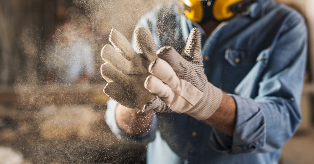 A bearded man wearing ear protection and work gloves clapping the gloves together, making dust puff out from them.