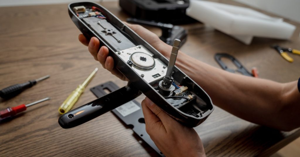 Hands holding a disassembled smart door lock handle exposing its internal circuity, with screwdrivers and tools nearby.